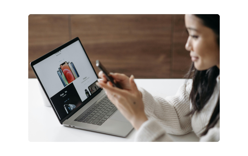 Woman browsing a dropshipping website on her phone while a laptop displays product listings.